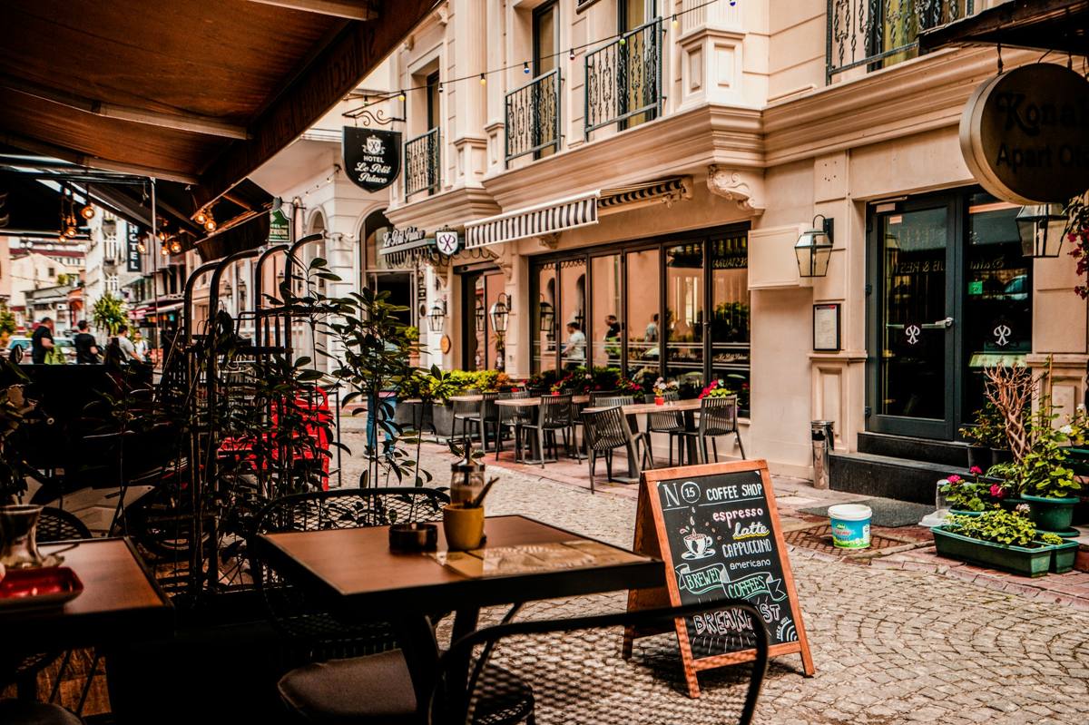 The shopfront on a cobbled side street: pavement tables under a canvas awning, potted geraniums along the kerb, and a chalkboard on the pavement listing espresso, latte and cappuccino.